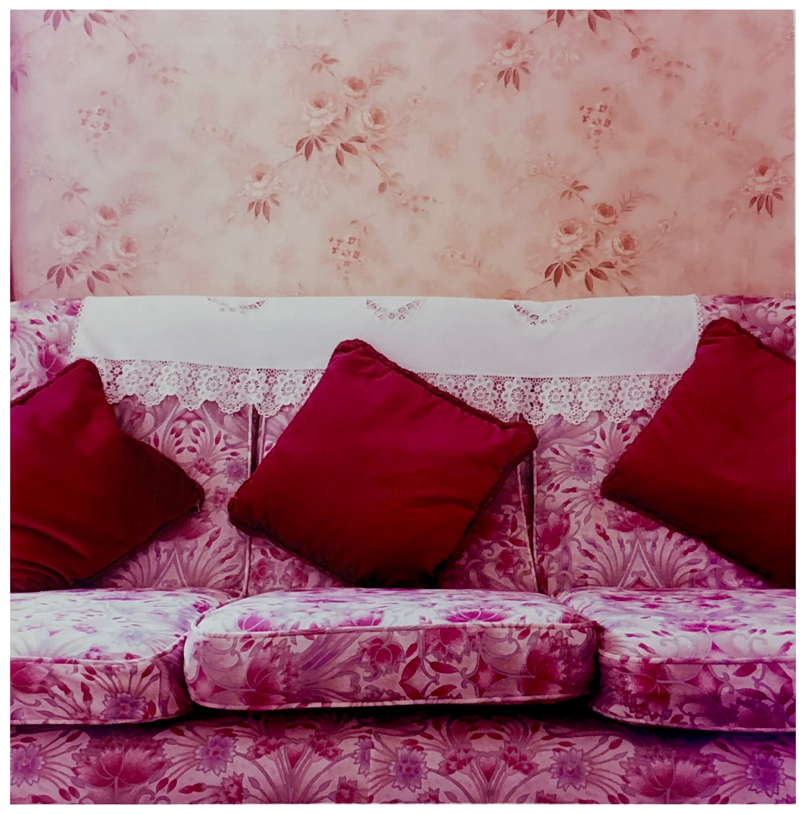 Photograph by Richard Heeps. A purple flowered sofa sits with three red cushions neatly placed and a crocheted antimacassar.  The background is a contrasting peach flowered wallpaper.  