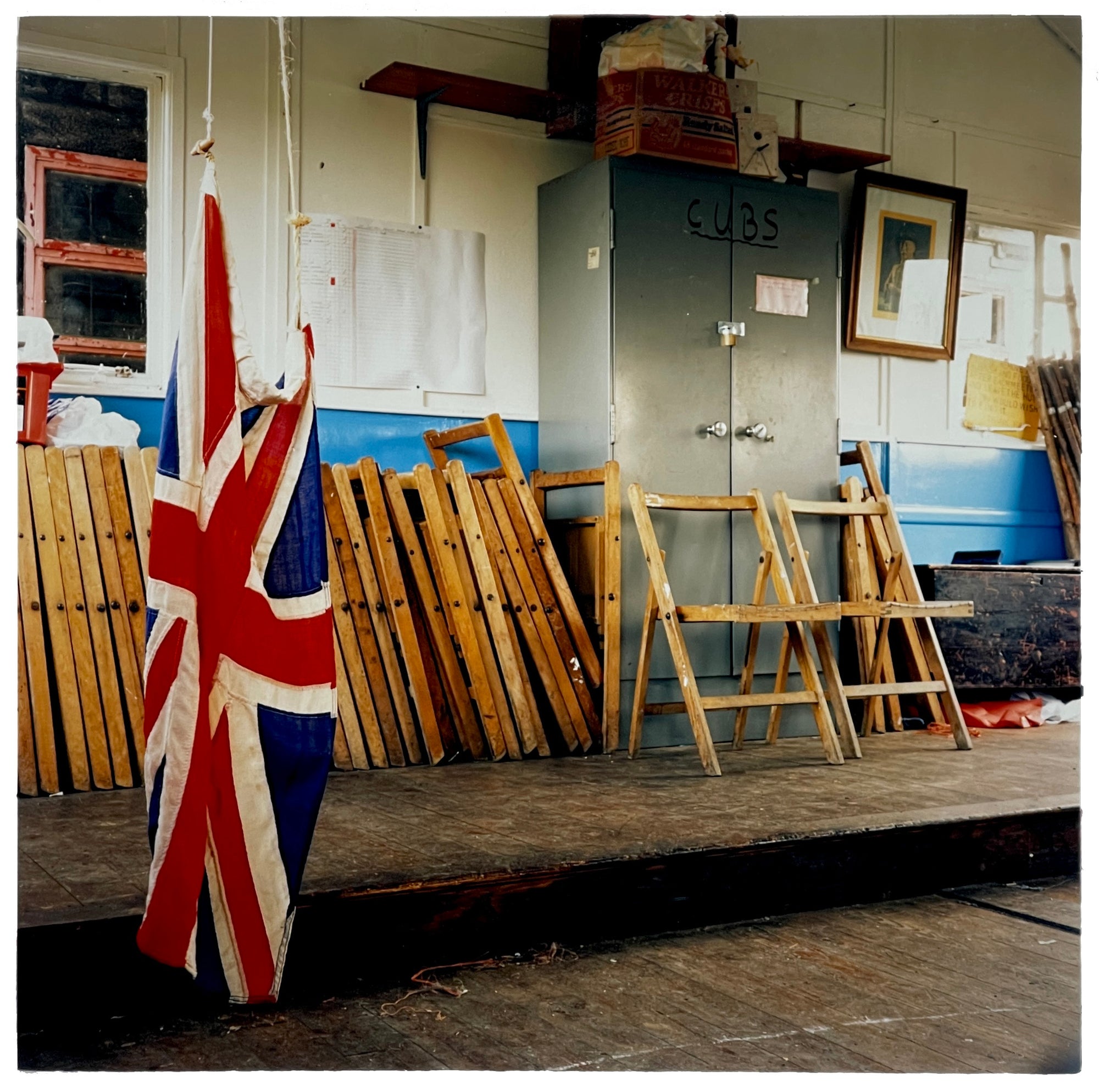 Photograph by Richard Heeps.  Inside a Cub Scout Hut, with the Union Jack Flag hanging and touching the floor.  The back half of the photo has a wooden platform with folding chairs on and the cubs own metal container.