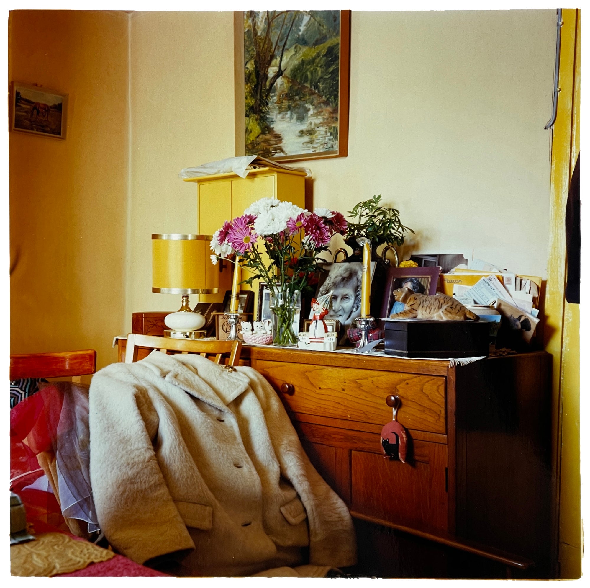 Photograph by Richard Heeps. A wooden dresser with photos, bunches of flowers, candles, ornaments, lamps and a full inbox on top.  In front of this busy piece of furniture are clothes laid on a chair, on top a white fur jacket. 