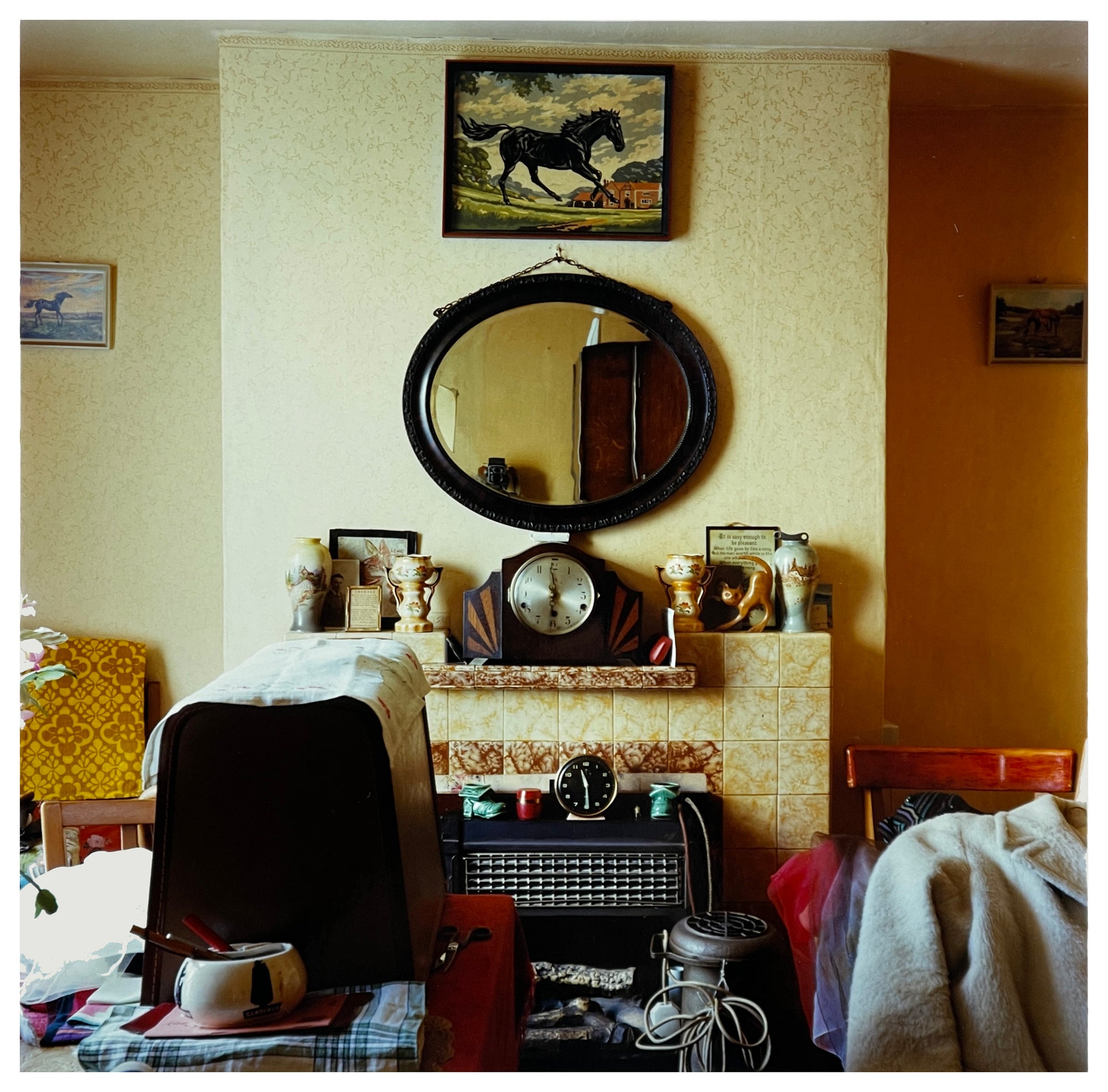 Photograph by Richard Heeps. A mantelpiece is in the middle of this photograph with an electric fire sitting in its hearth. The mantelpiece is surrounded and covered by paraphernalia. Above the mantelpiece is a mirror and above that a picture of a black horse. There is a boxed sewing machine in front of the mantelpiece.