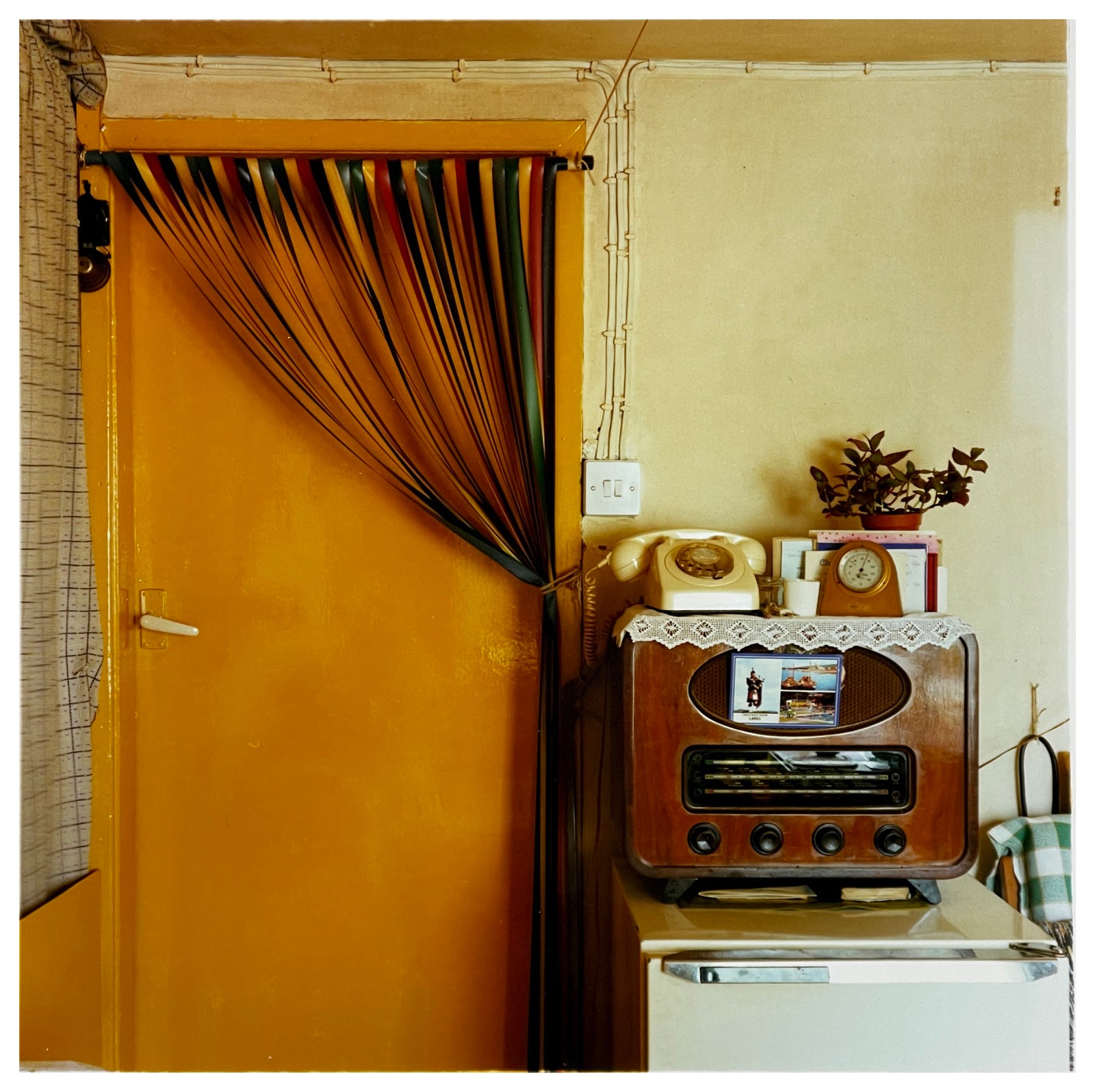 Photograph by Richard Heeps.  A vintage kitchen with an orange door and a plastic cream handle.  A door curtain is tied up over to the side of the door.  There sits to the right of the door a radio with a postcard from Largs tucked in the front.  On top of the radio is a doily on top of which sits a dial phone, carriage clock and houseplant sitting on top of the radio.