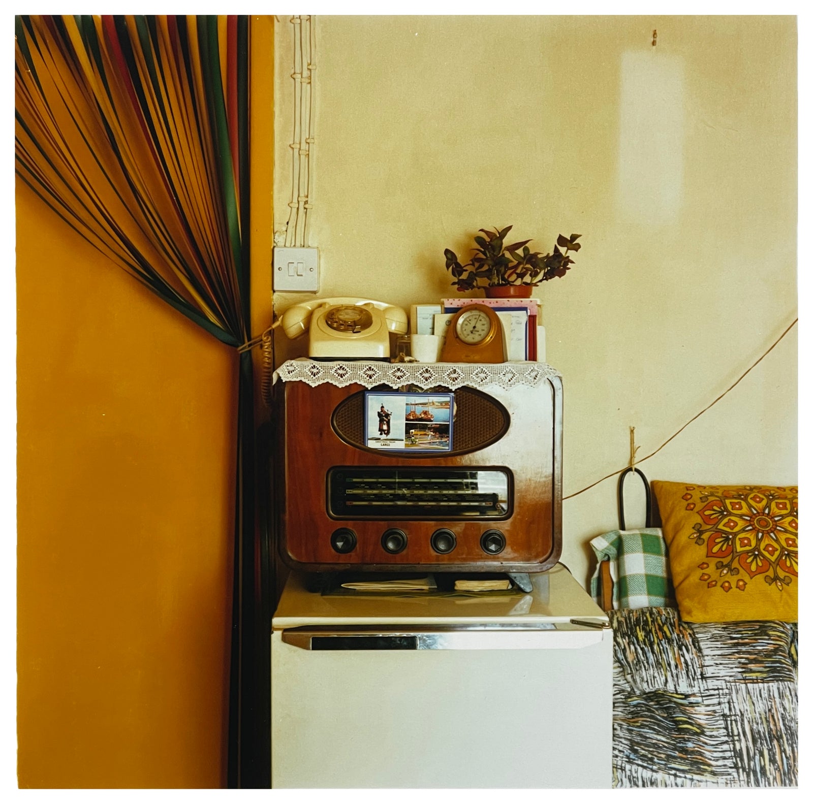 Photograph by Richard Heeps. This 1990s scene has a wooden-cased radio sitting on top of a fridge.  The radio is used as a shelf for a dial telephone, a pot plant and various other items. There is a tied-up door curtain to the left and cushions to the right.  