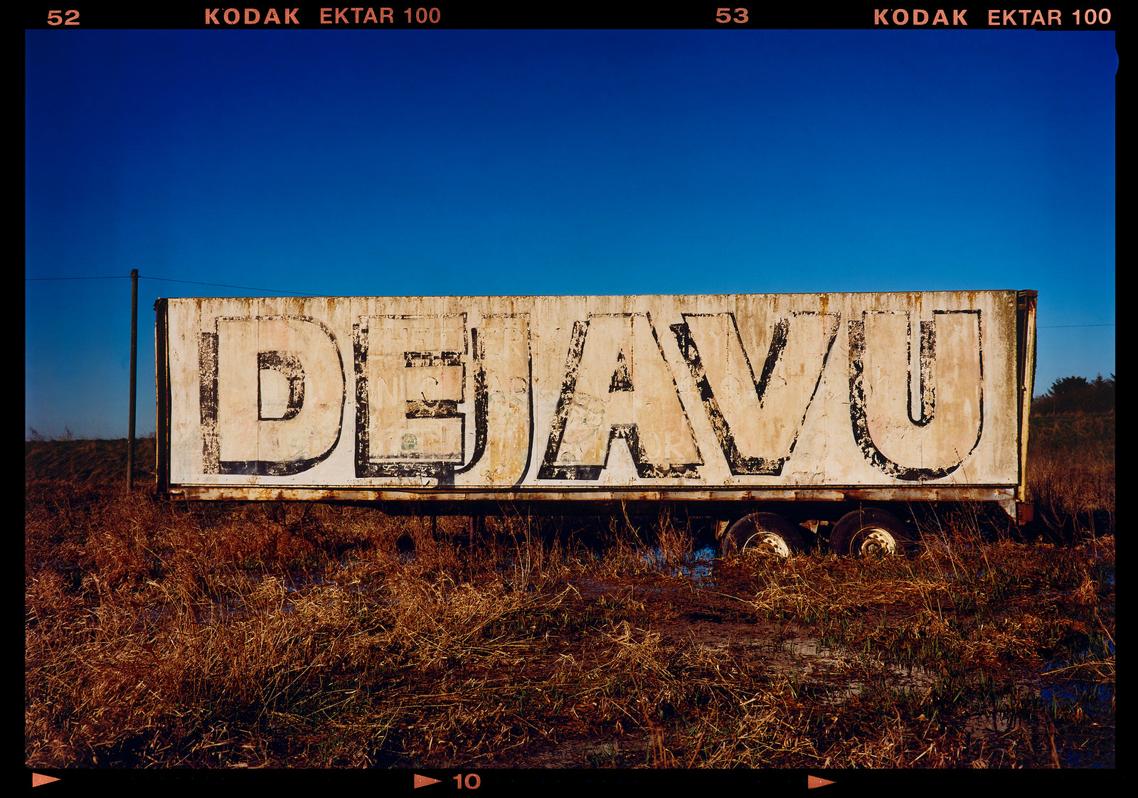 Photograph by Richard Heeps.  DEJAVU is painted on the side of a lorry trailer which sits in brown grass.  This is surrounded by a blue sky.