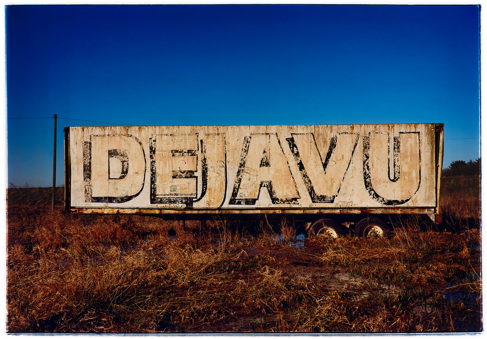 Photograph by Richard Heeps.  DEJAVU is painted on the side of a lorry trailer which sits in brown grass.  This is surrounded by a blue sky.