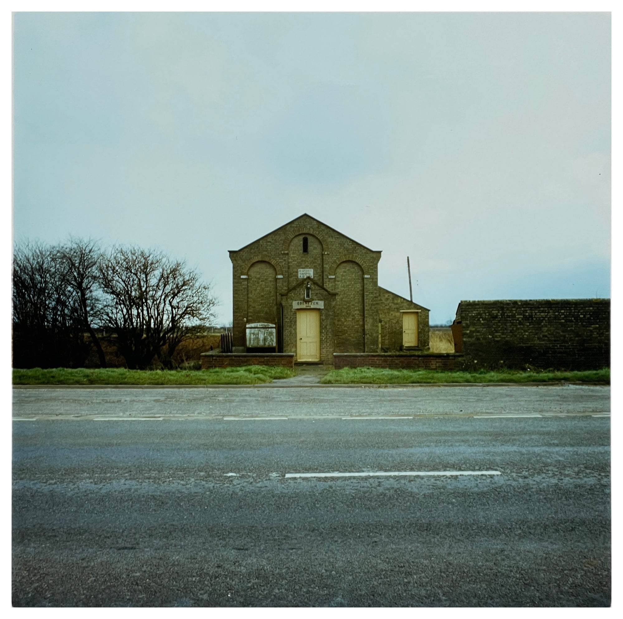 Photograph by Richard Heeps.  In the foreground is an empty road, on the opposite side of the road is a grass verge and then a yellow-doored, windowless, Chapel sitting solitary with no other buildings nearby.