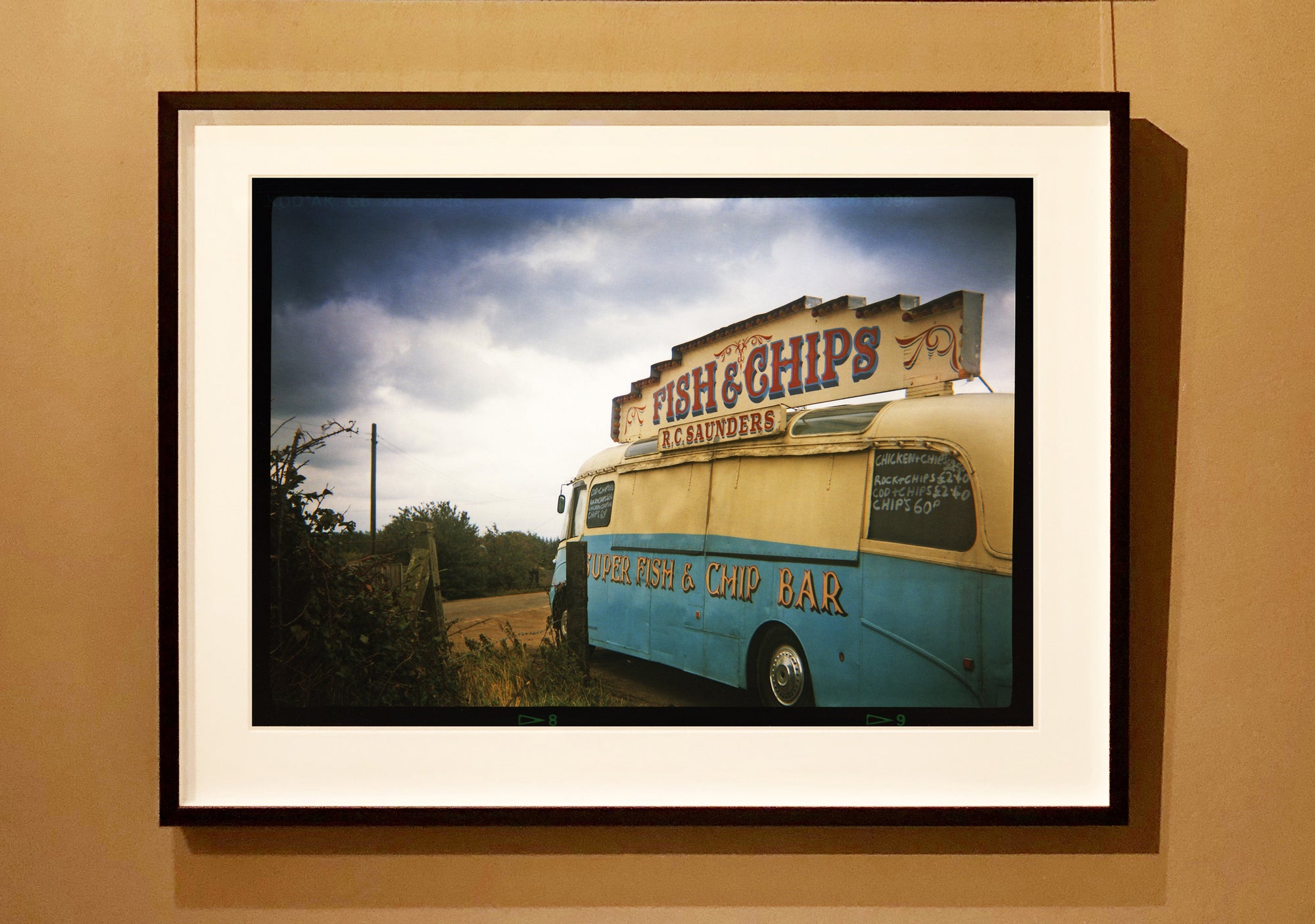 A moody Fenland sky is backdrop to a classic British Fish & Chips Van. The typography and the van itself has a fun fairground feel, contrary to the muted tones of this piece. Part of Richard Heeps series, 'A View of the Fens from the Car with Wings'.