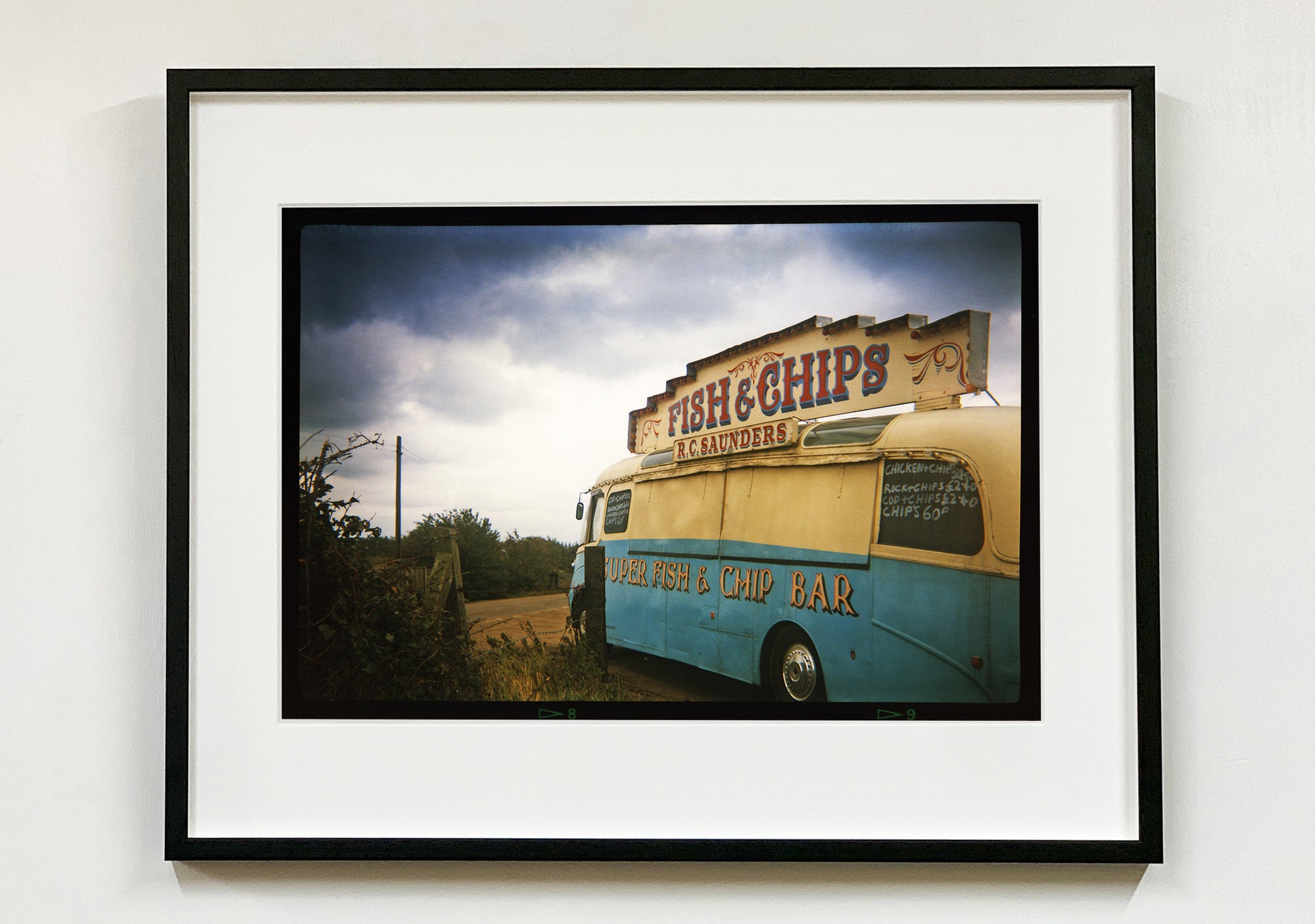 A moody Fenland sky is backdrop to a classic British Fish & Chips Van. The typography and the van itself has a fun fairground feel, contrary to the muted tones of this piece. Part of Richard Heeps series, 'A View of the Fens from the Car with Wings'.