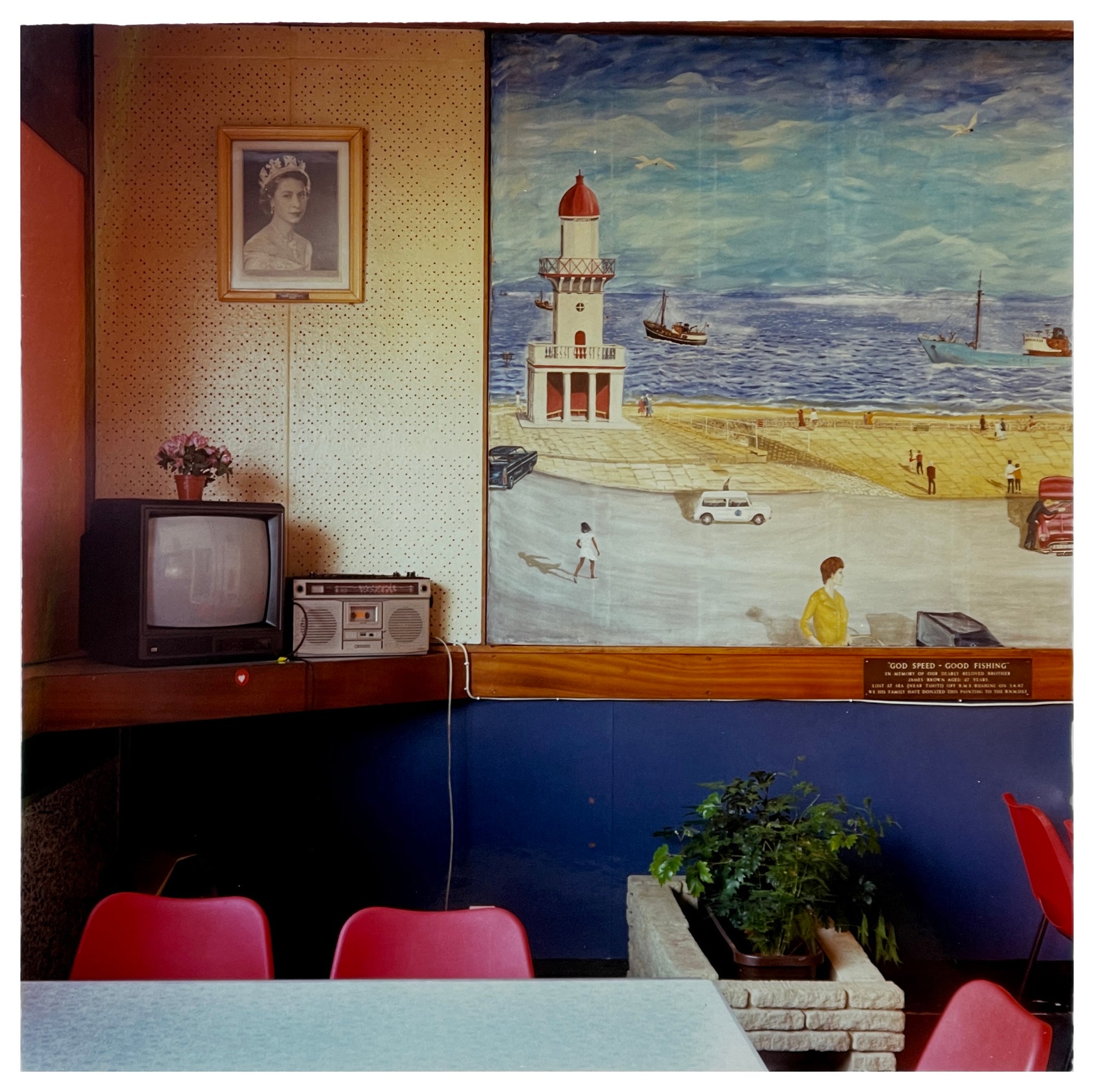 Photograph by Richard Heeps. This photo from 1986 is taken in a cafe which has a picture of Fleetwood beach erected In Memoriam which hangs next to a picture of Queen Elizabeth II.  Below is a shelf with a television and alongside that a radio.  In the room are cafe-style plastic seating and chairs.  