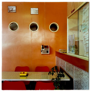 Photograph by Richard Heeps.  Taken in the Fisherman's Mission cafe, on the wall are three portholes in an orange-coloured wooden wall.  On the wall is a payphone and this part of the room holds a cafe-style table and chairs, on the table is a yellow ashtray.