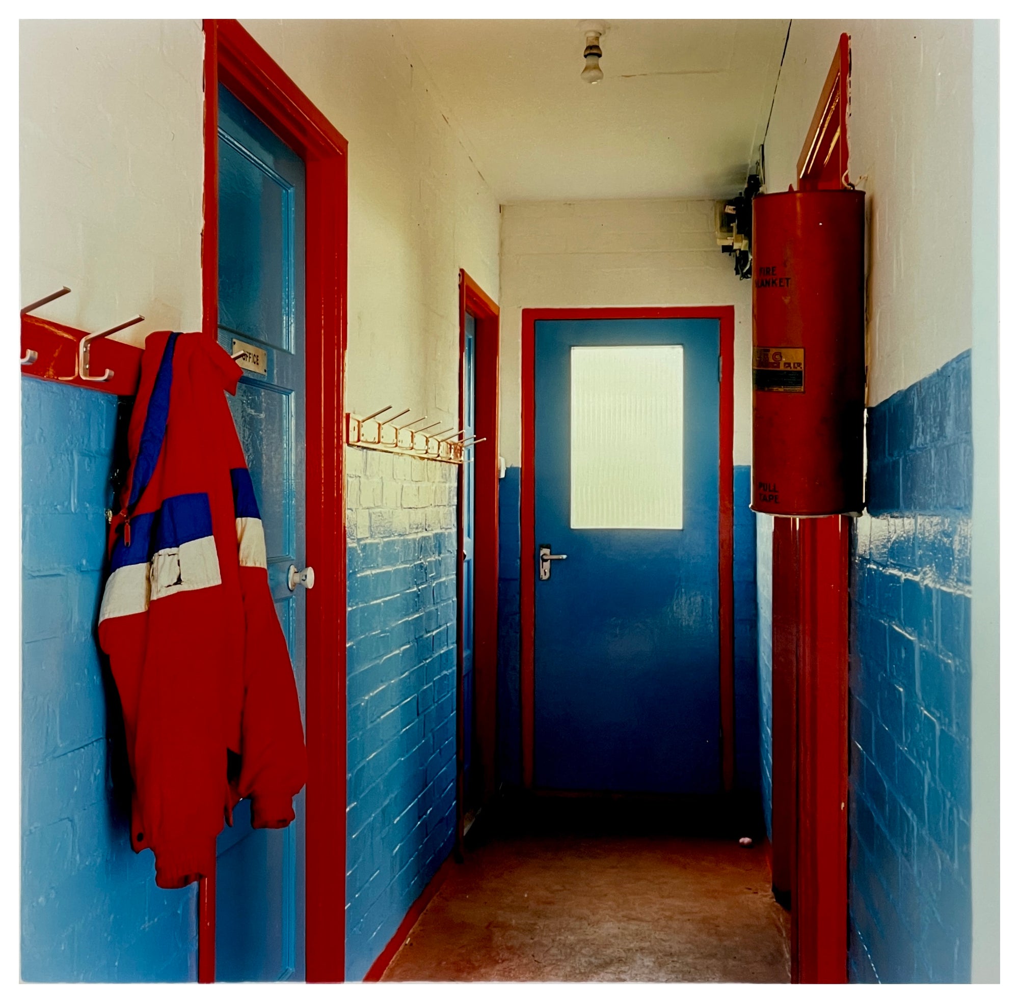 Photograph by Richard Heeps.  A hallway in a scouts hut with painted walls, the top half of the walls are painted cream and the bottom are painted blue.  There are two blue painted doors which have a red door frame.  There are red painted coat hooks on the left hand side on the wall on which is hanging a jacket made up of nearly identical red and blue colours.