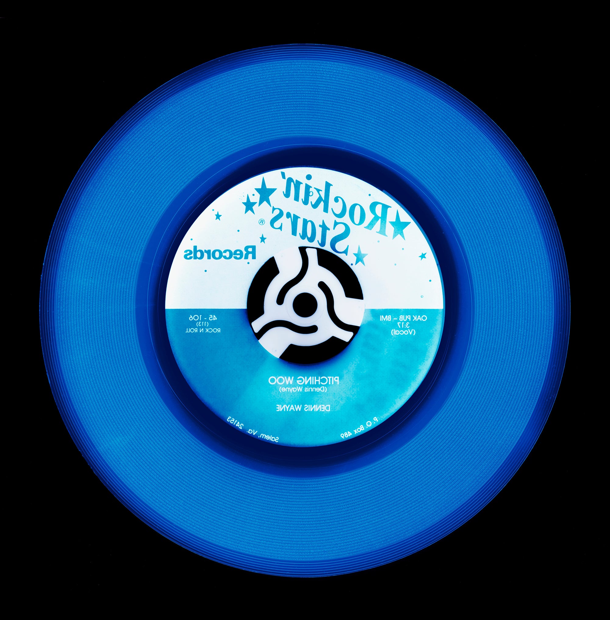 Photograph by Natasha Heidler and Richard Heeps.  A denim blue record with darker blue grooves and the record label is half white, half blue with writing in the opposite blue and white.  This vinyl sits on a black background.