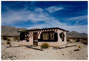 Photograph by Richard Heeps. An abandoned building sitting alone in a desert, tumble weed on the ground and hills in the background. A blue sky with white clouds.