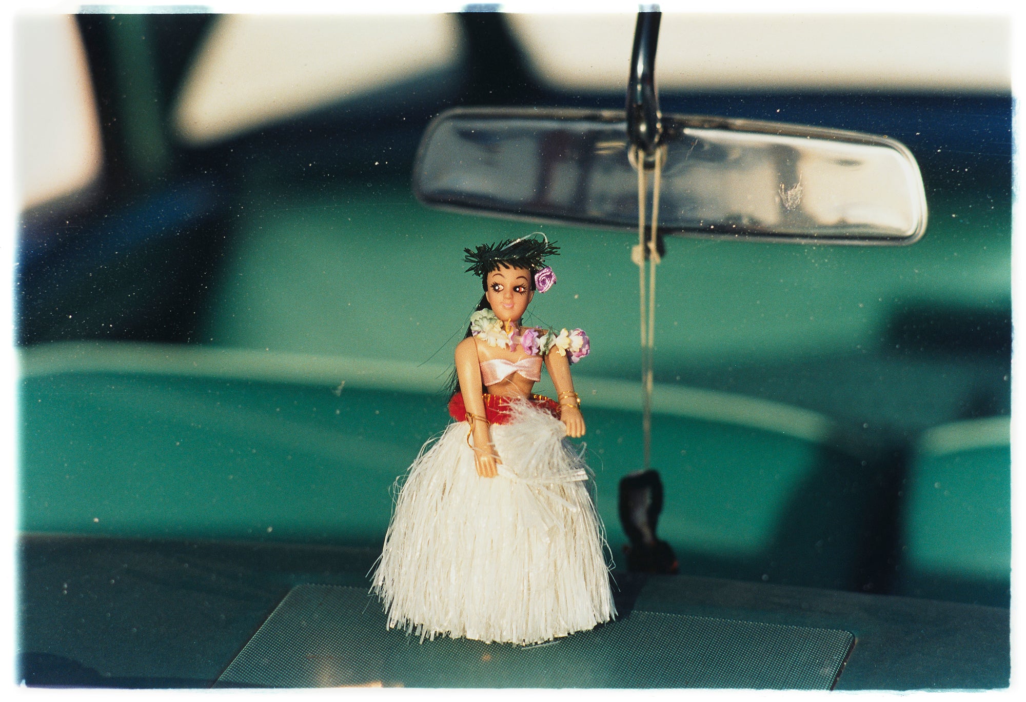 Photograph by Richard Heeps.  A hula doll sits on a dashboard, below the rear view mirror.  The car's interior is green and out of focus.