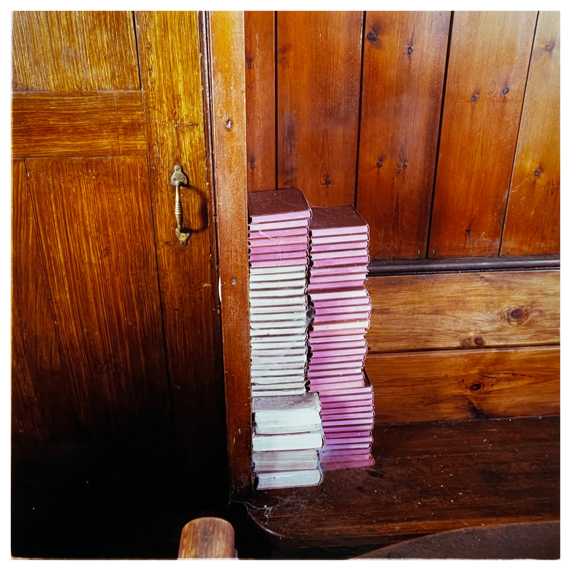 Photograph by Richard Heeps.  Stacked up in the corner of a wooden pew next to a wooden door is two columns of hymn books.  The hymn books are red hard back some with white paper edging and some with pink.