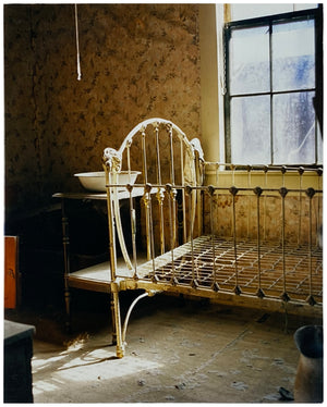 Photograph by Richard Heeps. A run down now unused room with the metal surround of a cot bed and no mattress, at the end of the bed a wash stand with a bowl on top. Light shines in the room from the window.