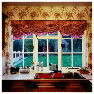 Photograph by Richard Heeps. A kitchen sink from 1989 with brown taps and bowl, and surrounded by three bottles of washing up liquid.  The window above the sink looks out on a neat garden and is topped with orange ruched curtains.  The wallpaper has a sequence of orange flowers linked with orange ribbon.  