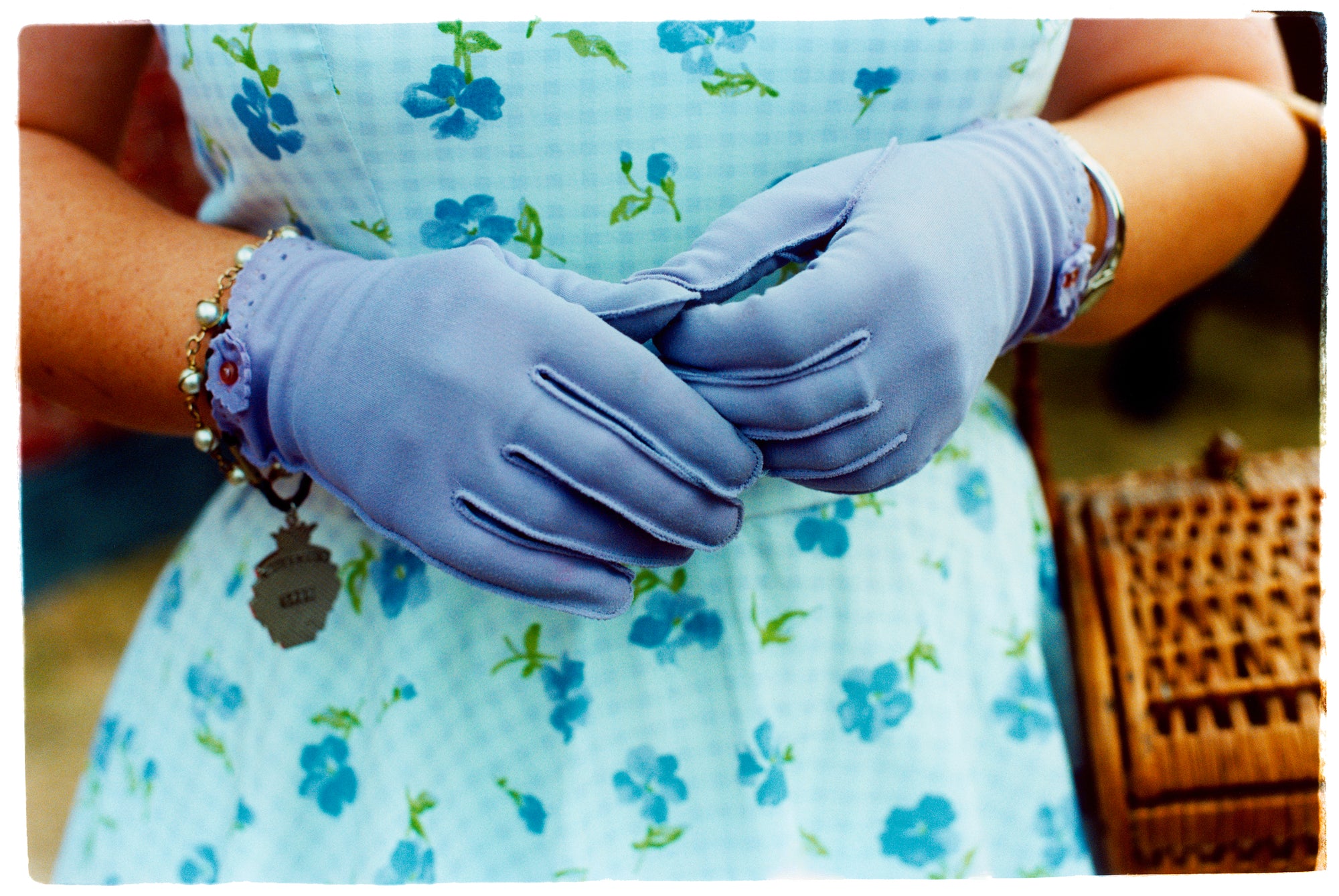 Photograph by Richard Heeps.  Retro lilac gloves are together in front of a blue flowered dress worn by a woman at the Goodwood Revival.  To the side is a wicker basket.