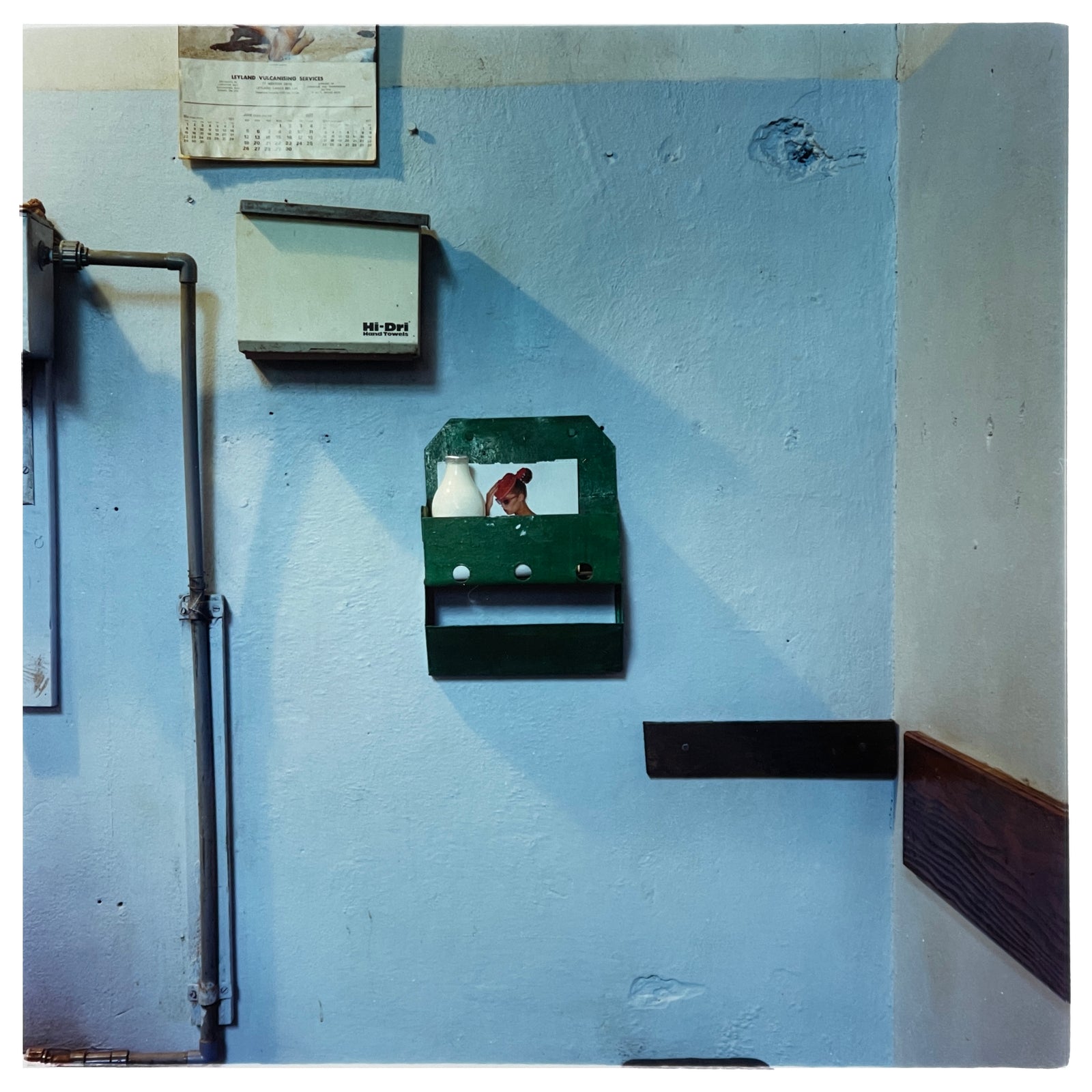 Photograph by Richard Heeps. A light blue factory wall with a green milk rack at its centre, with a glass pint of milk and a photo of a woman attached to the back of the milk rack.