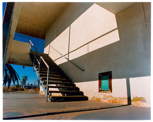 Photograph by Richard Heeps. The photograph has a metal staircase on the outside of a cream colour motel. The staircase has a ceiling but no sides so leads to blue sky. Behind the building is a blue sky and palm trees of the Californian Desert.