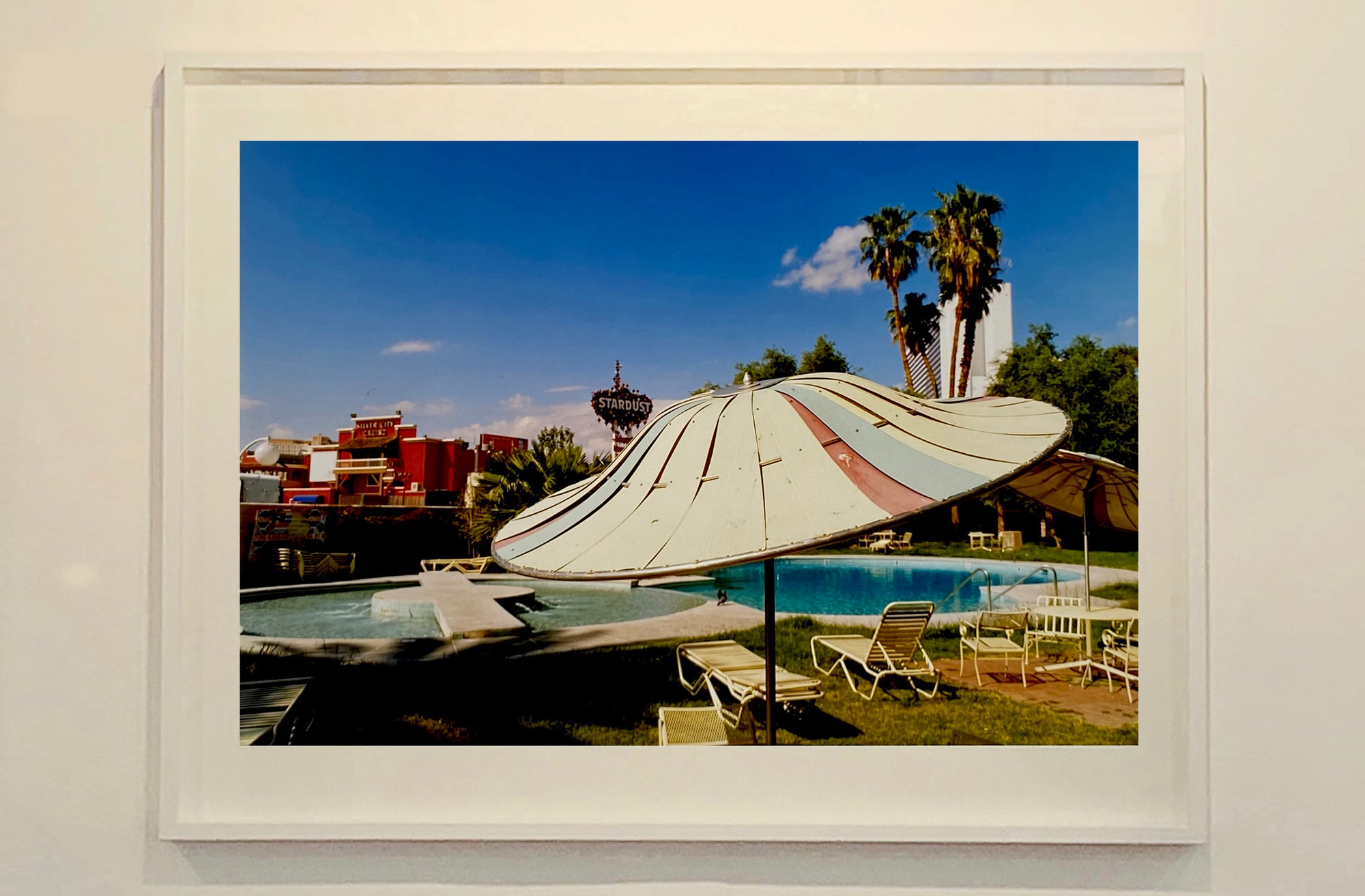 A vintage parasol, poolside at the El Morocco Motel, Las Vegas, Nevada, overlooked by palm trees, blue skies and iconic neon signs. Part of Richard Heeps' 'Dream in Colour' series.
