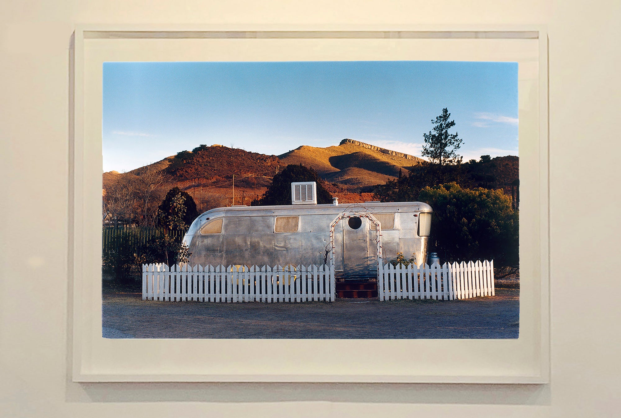 This artwork brings about a romantic dream of living in an RV. Captured in the morning sun at the start of a beautiful Arizona day, this RV is surrounded by both a white picket fence and a beautiful landscape, with the sun tickling the hilltops. Photographed as part of Richard Heeps' 'Dream in Colour' series.
