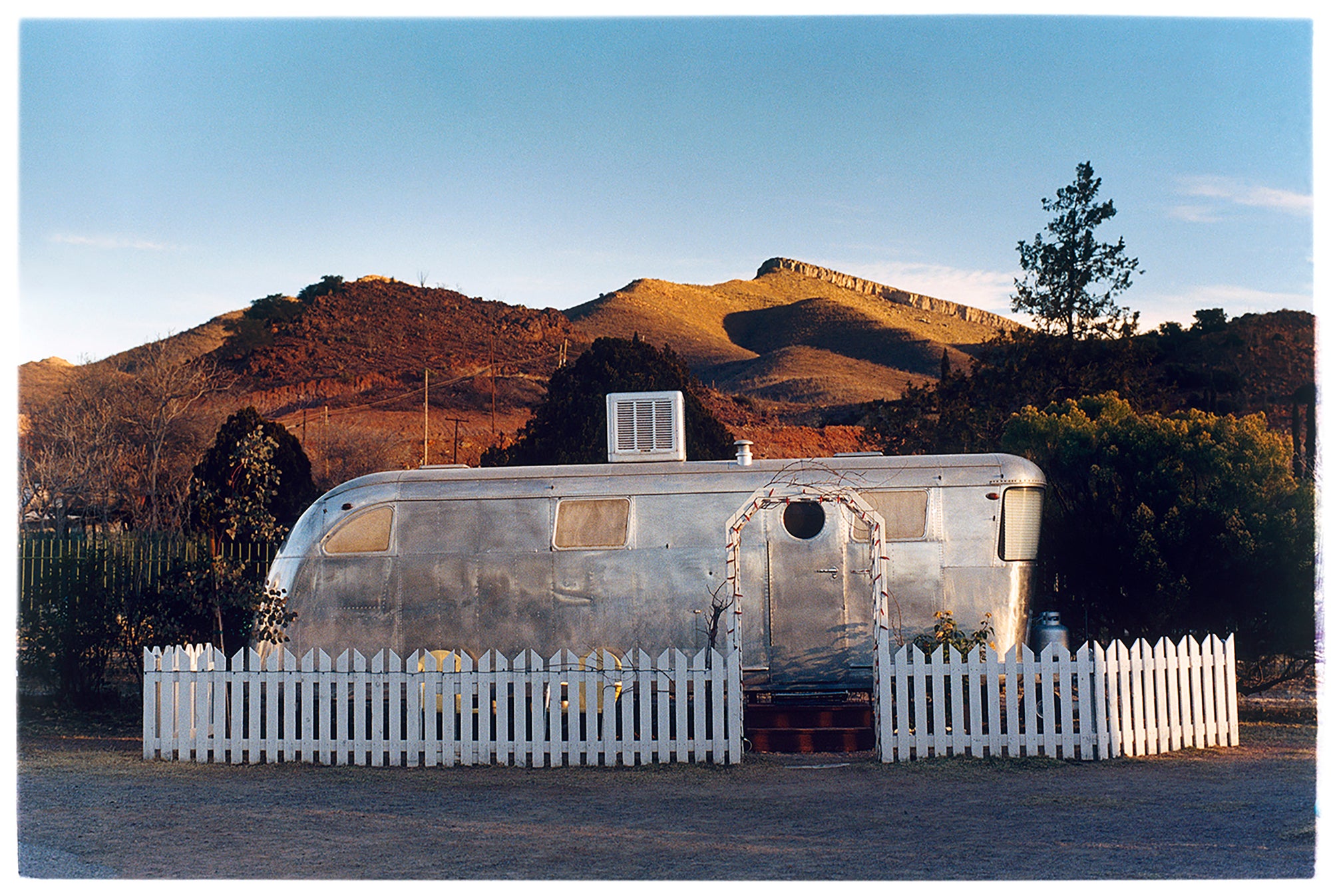 This artwork brings about a romantic dream of living in an RV. Captured in the morning sun at the start of a beautiful Arizona day, this RV is surrounded by both a white picket fence and a beautiful landscape, with the sun tickling the hilltops. Photographed as part of Richard Heeps' 'Dream in Colour' series.