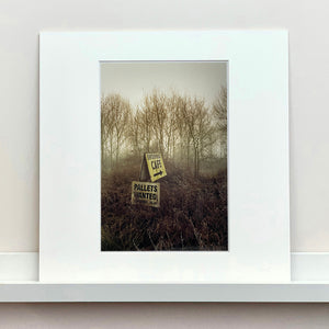 Mounted photograph by Richard Heeps.  A roadside sign for The Enterprise Cafe and Pallets Wanted sit in brown shrub with wintry trees and sky in the background.