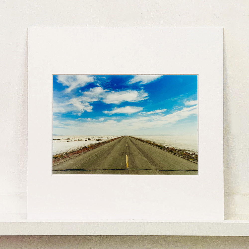 Photograph by Richard Heeps.  In the centre of the empty approach road going off into the distance, blue skies and white clouds fill the sky and either side of the road the flat salt plains.