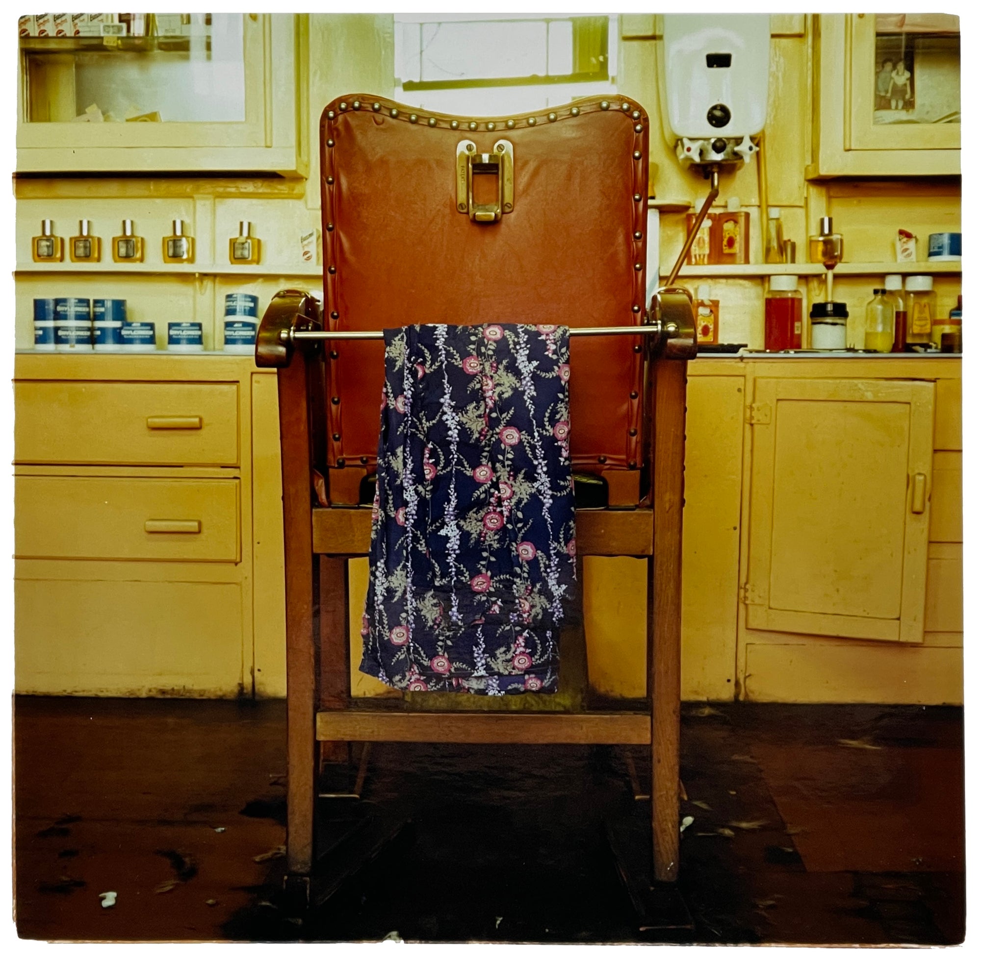 Photograph by Richard Heeps. An antique barbers chair, with a customer robe draped over the front, sits in front of an aged yellow side board on which sits many barber type lotions. There is a water heater sitting on the wall above the sideboard