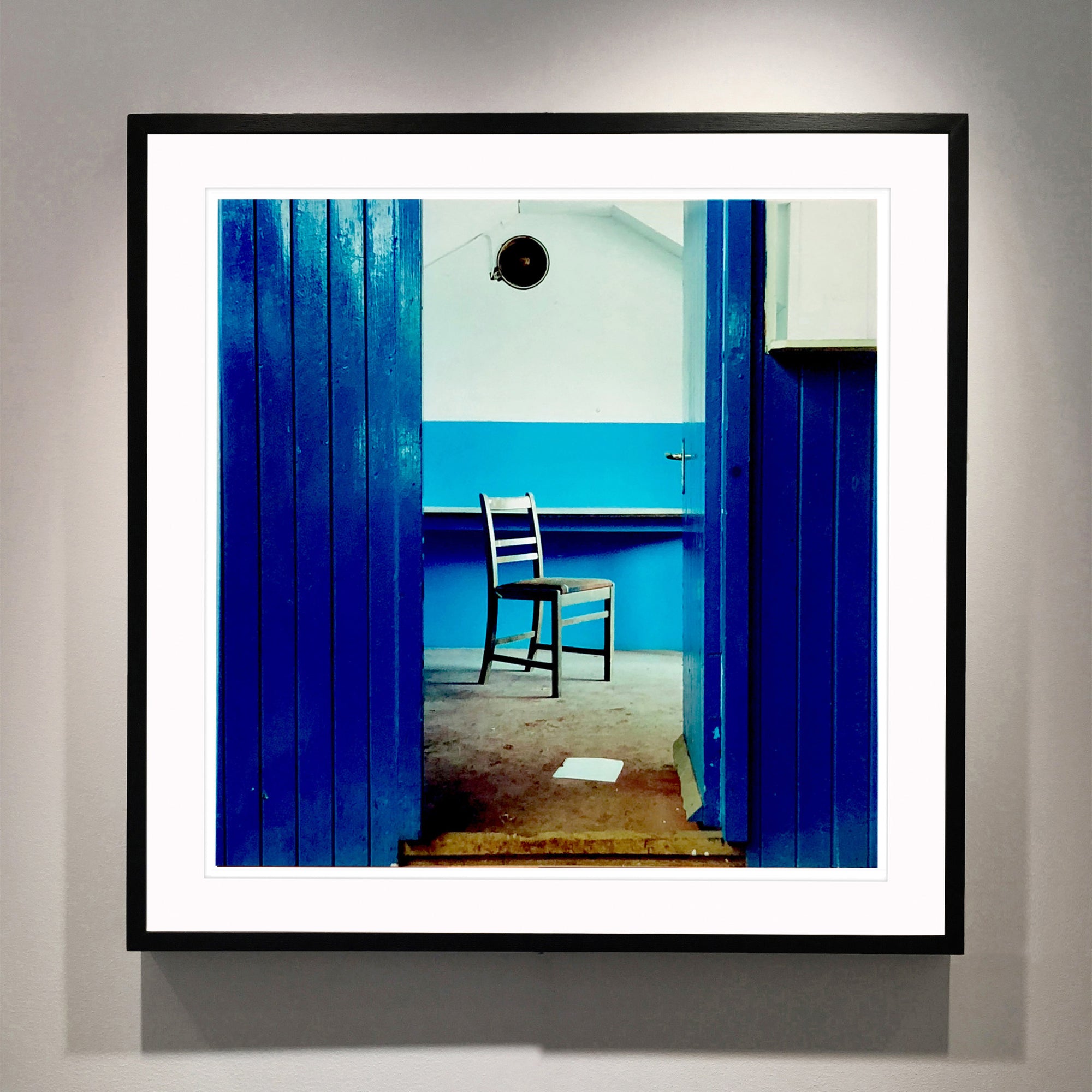 A photograph of a chair which sits in a derelict factory in a colour block blue painted room, the perspective creates depth.