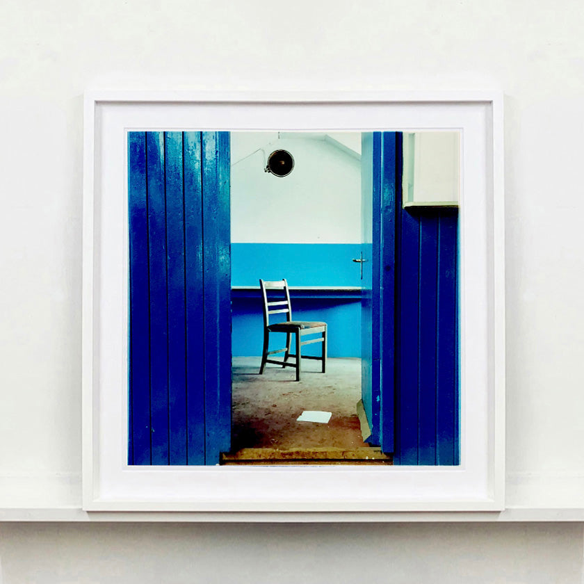 A photo of a chair which sits in a derelict factory in a colour block blue painted room, the perspective creates depth.