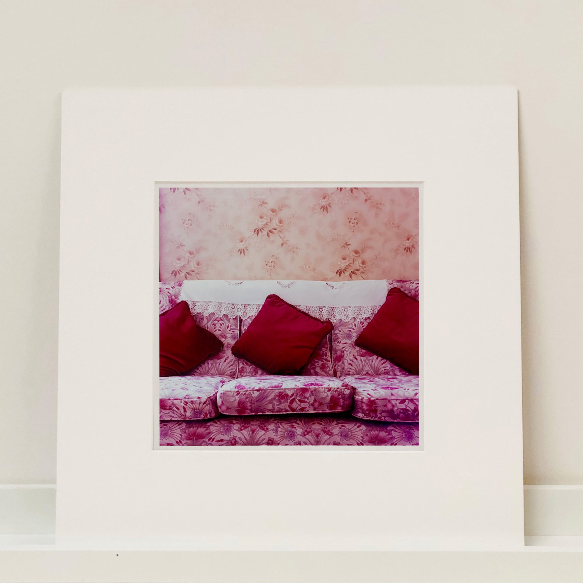 Photograph by Richard Heeps. A purple flowered sofa sits with three red cushions neatly placed and a crocheted antimacassar.  The background is a contrasting peach flowered wallpaper.  