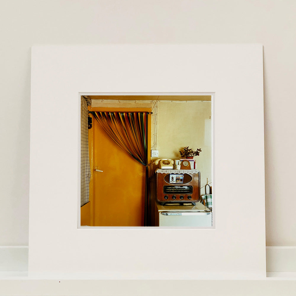 Photograph by Richard Heeps.  A vintage kitchen with an orange door and a plastic cream handle.  A door curtain is tied up over to the side of the door.  There sits to the right of the door a radio with a postcard from Largs tucked in the front.  On top of the radio is a doily on top of which sits a dial phone, carriage clock and houseplant sitting on top of the radio.