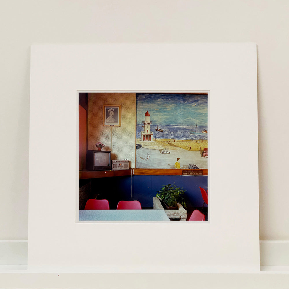 Photograph by Richard Heeps. This photo from 1986 is taken in a cafe which has a picture of Fleetwood beach erected In Memoriam which hangs next to a picture of Queen Elizabeth II.  Below is a shelf with a television and alongside that a radio.  In the room are cafe-style plastic seating and chairs.  