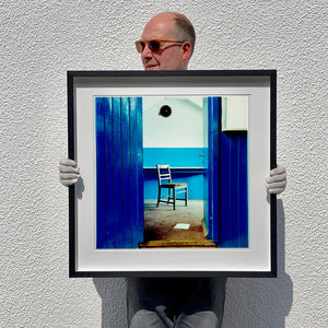 A framed photograph of a chair which sits in a derelict factory in a colour block blue painted room, the perspective creates depth.