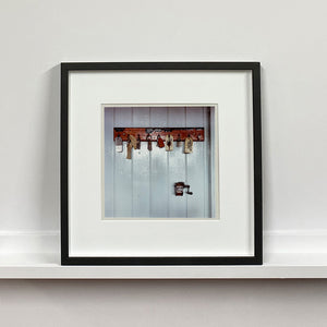 A vintage photograph of a dark wooden key holder carrying a selection of keys and fobs.  The holder is fixed on a white-painted wooden slated wall.  There is a pencil sharpener attached to the wall also, below the key holder.  Photograph by Richard Heeps. Browser Black Frame Square.