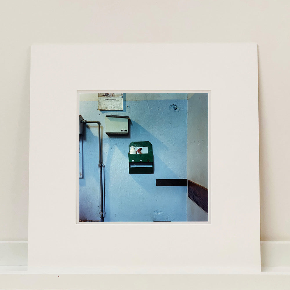 Photograph by Richard Heeps. A light blue factory wall with a green milk rack at its centre, with a glass pint of milk and a photo of a woman attached to the back of the milk rack.