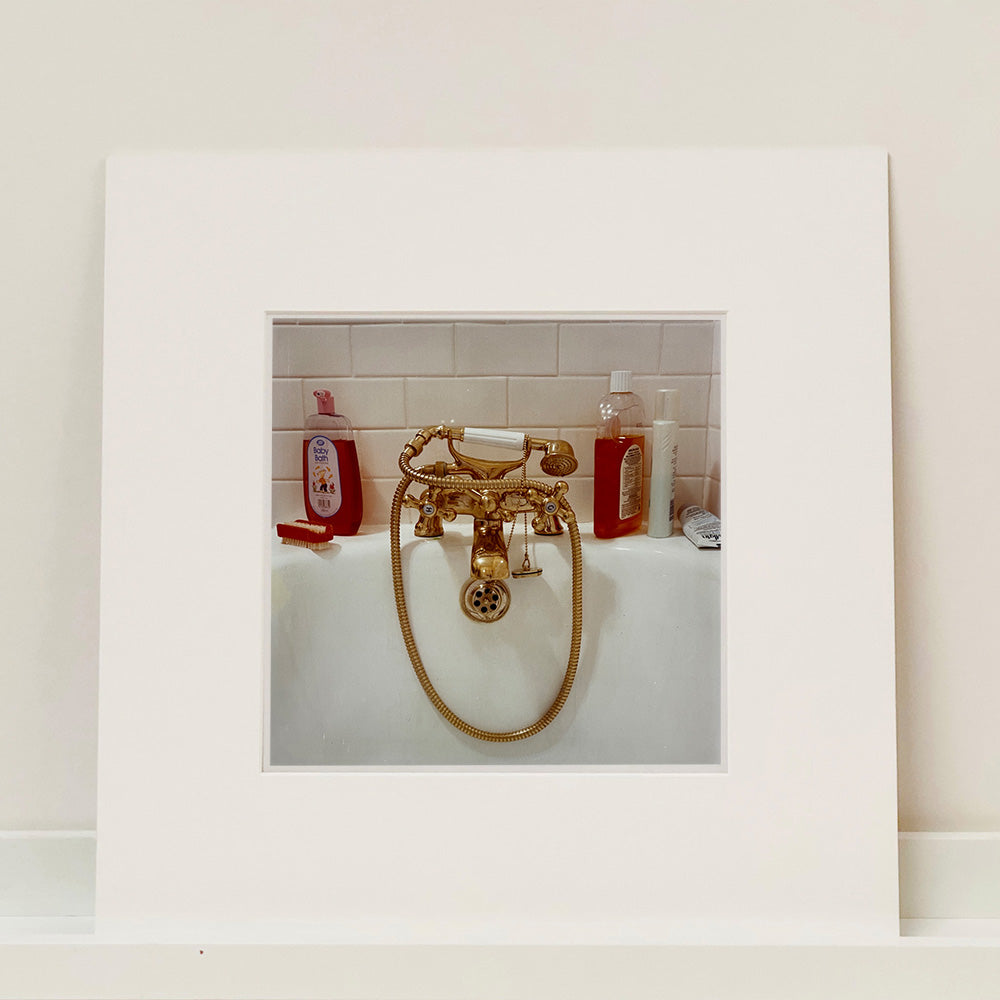 Photograph by Richard Heeps.  Gold taps and hand held shower at the end of a bath.  Red toned bottles of shampoo and baby bath contrast against the white bath and white bathroom tiles.