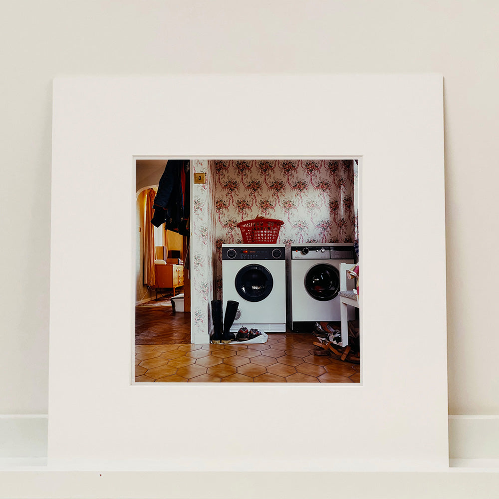 Photograph by Richard Heeps.  A 1980s family utility room with a washing machine, a tumble dryer, brown linoleum and flowery wallpaper.