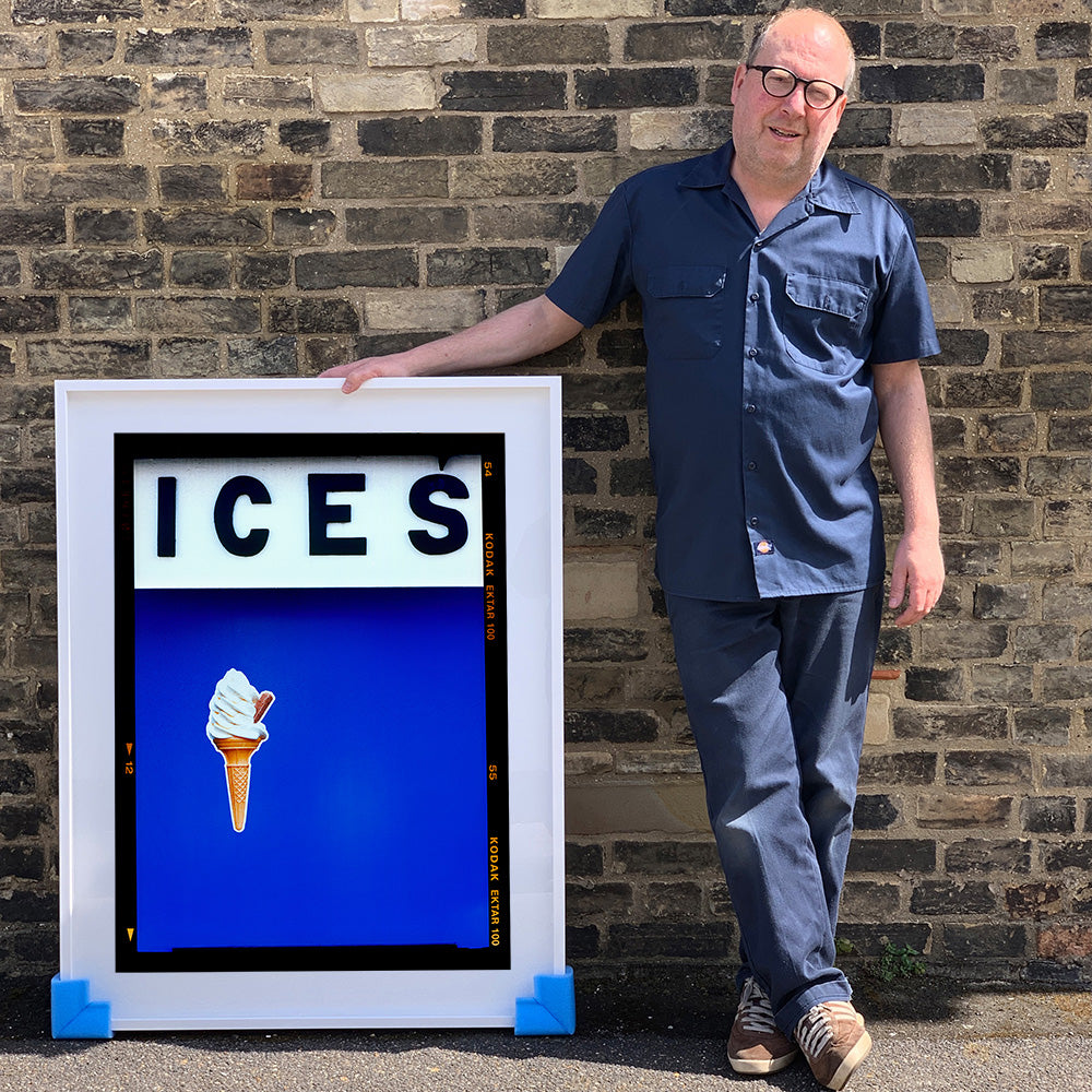 Photograph by Richard Heeps.  Richard Heeps holds a white framed print. At the top of the print, black letters spell out ICES and below is depicted a 99 icecream cone sitting left of centre against a blue coloured background.  