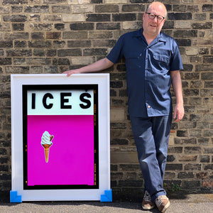 Photograph by Richard Heeps.  Richard Heeps holds a white framed print. At the top of the print, black letters spell out ICES and below is depicted a 99 icecream cone sitting left of centre against a pink coloured background.  