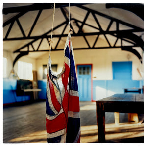 Photograph by Richard Heeps.  A Union Jack hangs from the rafters of a Scout Hut.  The room has blue doors, a wooden floor, an old wooden table and wooden rafters. 
