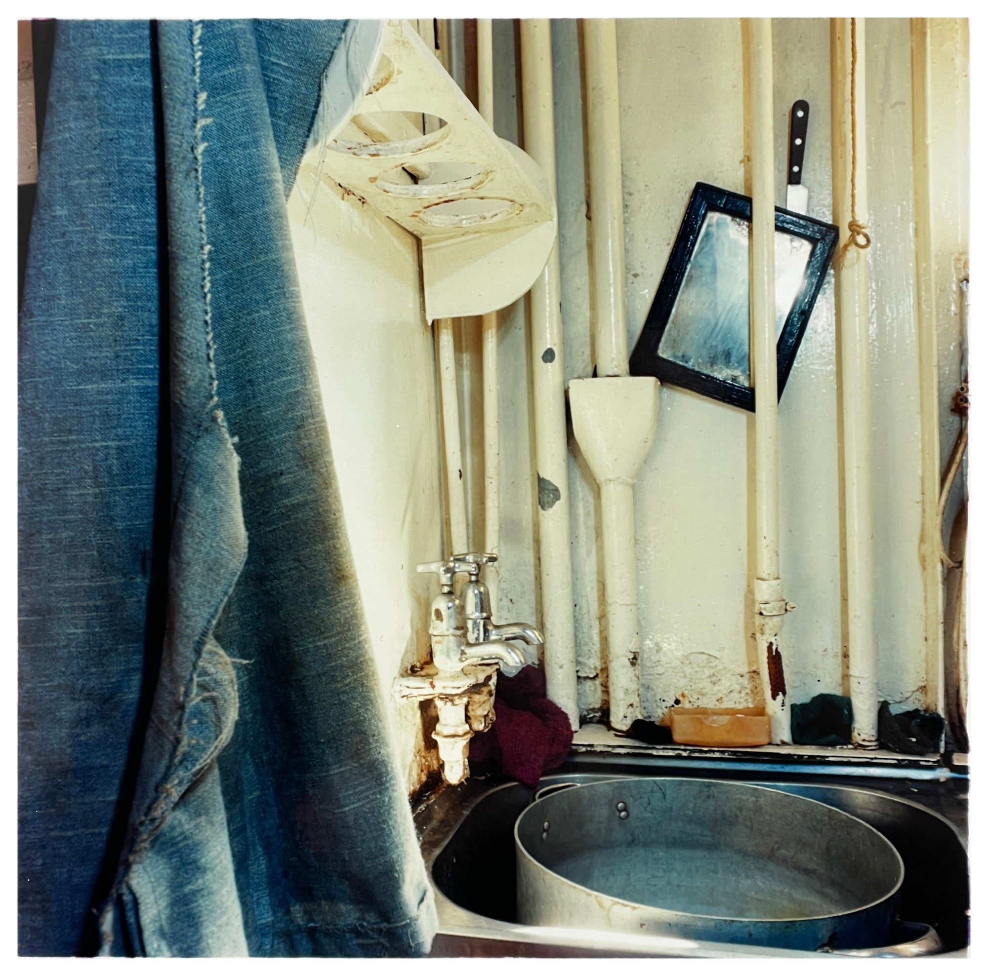 Photograph by Richard Heeps.  Small metal sink sits with a metal bowl.  The walls are covered with an old cream paint, which has chips and shows signs of decay.  A denim shirt hangs on the left hand side.