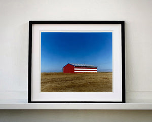 'Stars & Stripes Barn' shows a depiction of the American flag painted on this building in Oakhurst, California. It sits on the horizon against a vast blue sky. This artwork is part of Richard Heeps' 'Dream in Colour' series.