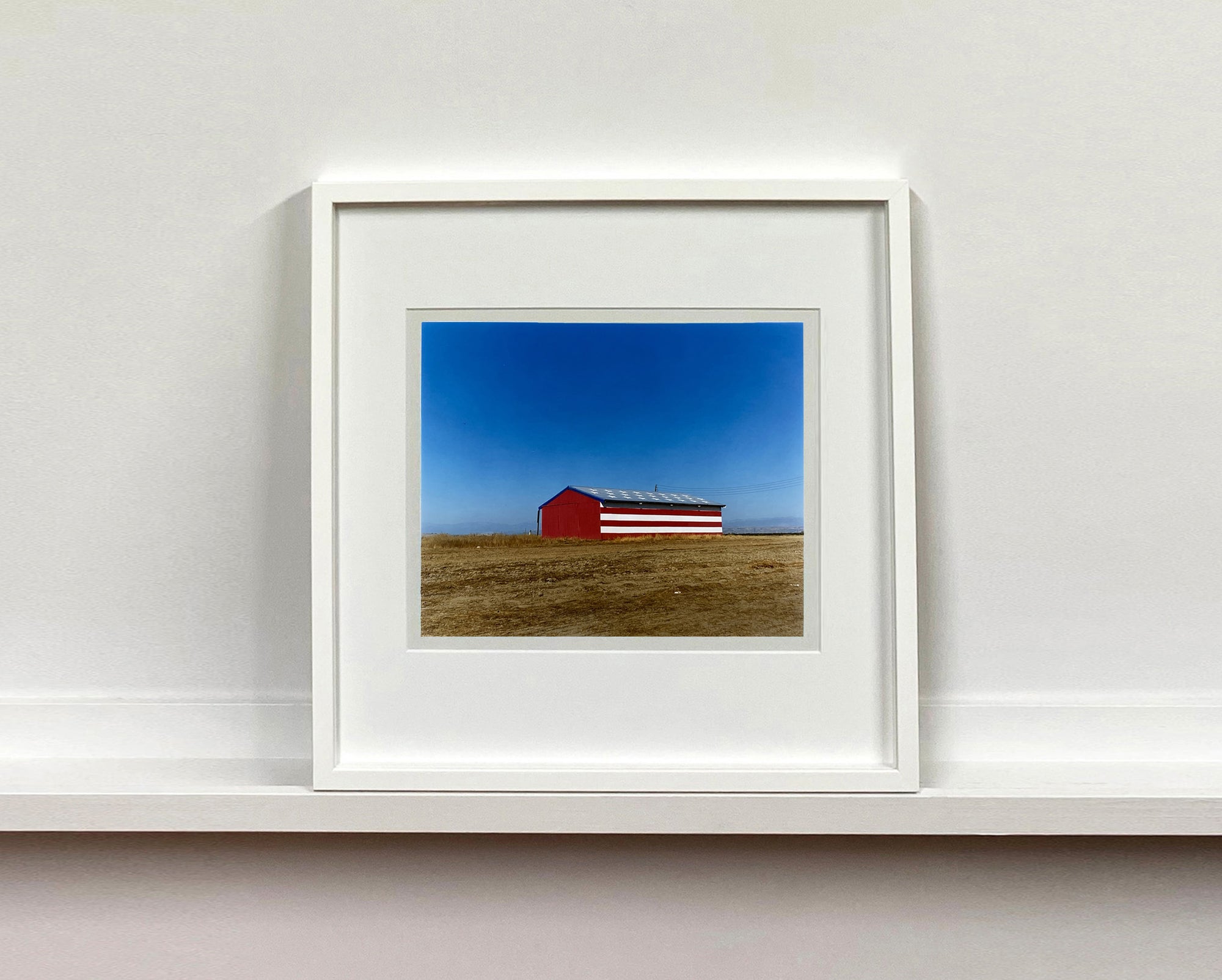 'Stars & Stripes Barn' shows a depiction of the American flag painted on this building in Oakhurst, California. It sits on the horizon against a vast blue sky. This artwork is part of Richard Heeps' 'Dream in Colour' series.