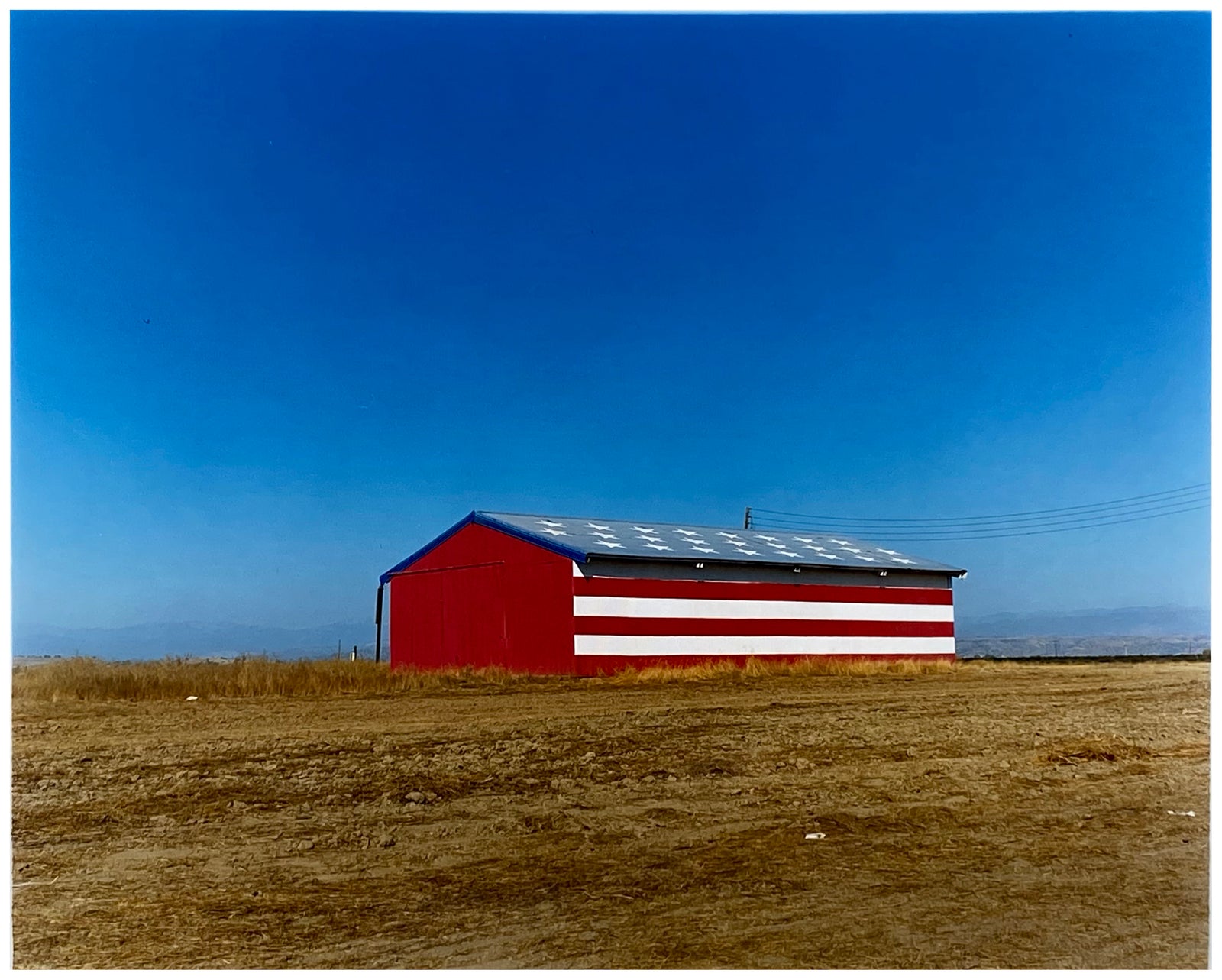 'Stars & Stripes Barn' shows a depiction of the American flag painted on this building in Oakhurst, California. It sits on the horizon against a vast blue sky. This artwork is part of Richard Heeps' 'Dream in Colour' series.