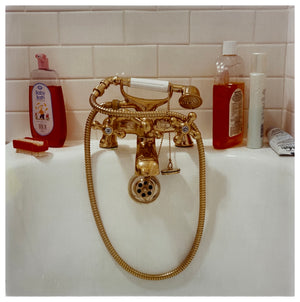 Photograph by Richard Heeps.  Gold taps and hand held shower at the end of a bath.  Red toned bottles of shampoo and baby bath contrast against the white bath and white bathroom tiles.
