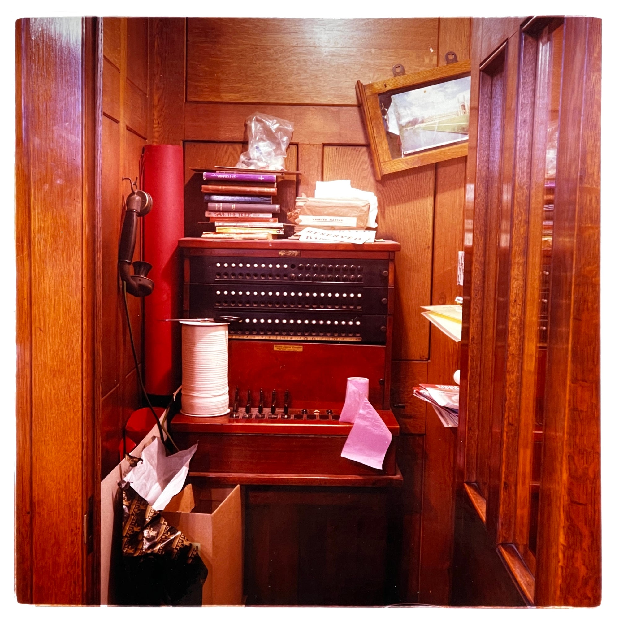 Photograph by Richard Heeps.  A wood panelled booth with an antique phone handset hanging on a hook on the left and an exchange in the middle, piled with books.