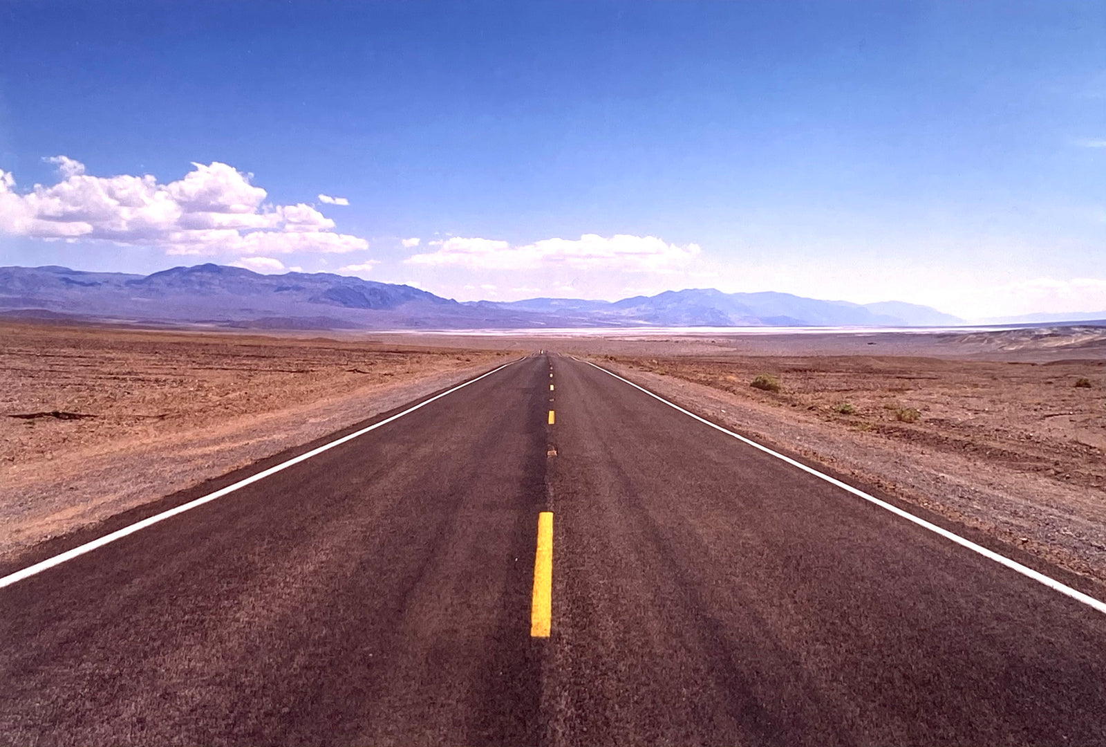 'The Road to Death Valley', taken in the Majova Desert, California, features a clouded blue sky met my mountains on the horizon. This American landscape artwork is part of Richard Heeps' 'Dream in Colour' series.