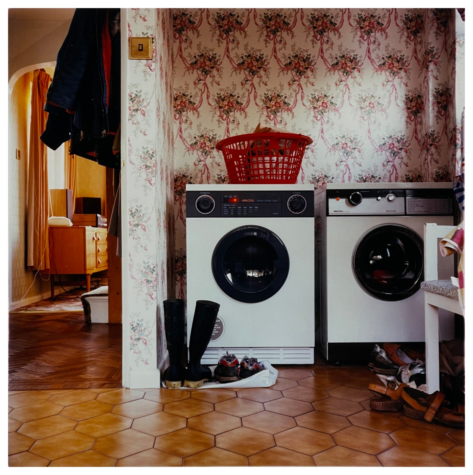 Photograph by Richard Heeps.  A 1980s family utility room with a washing machine, a tumble dryer, brown linoleum and flowery wallpaper.